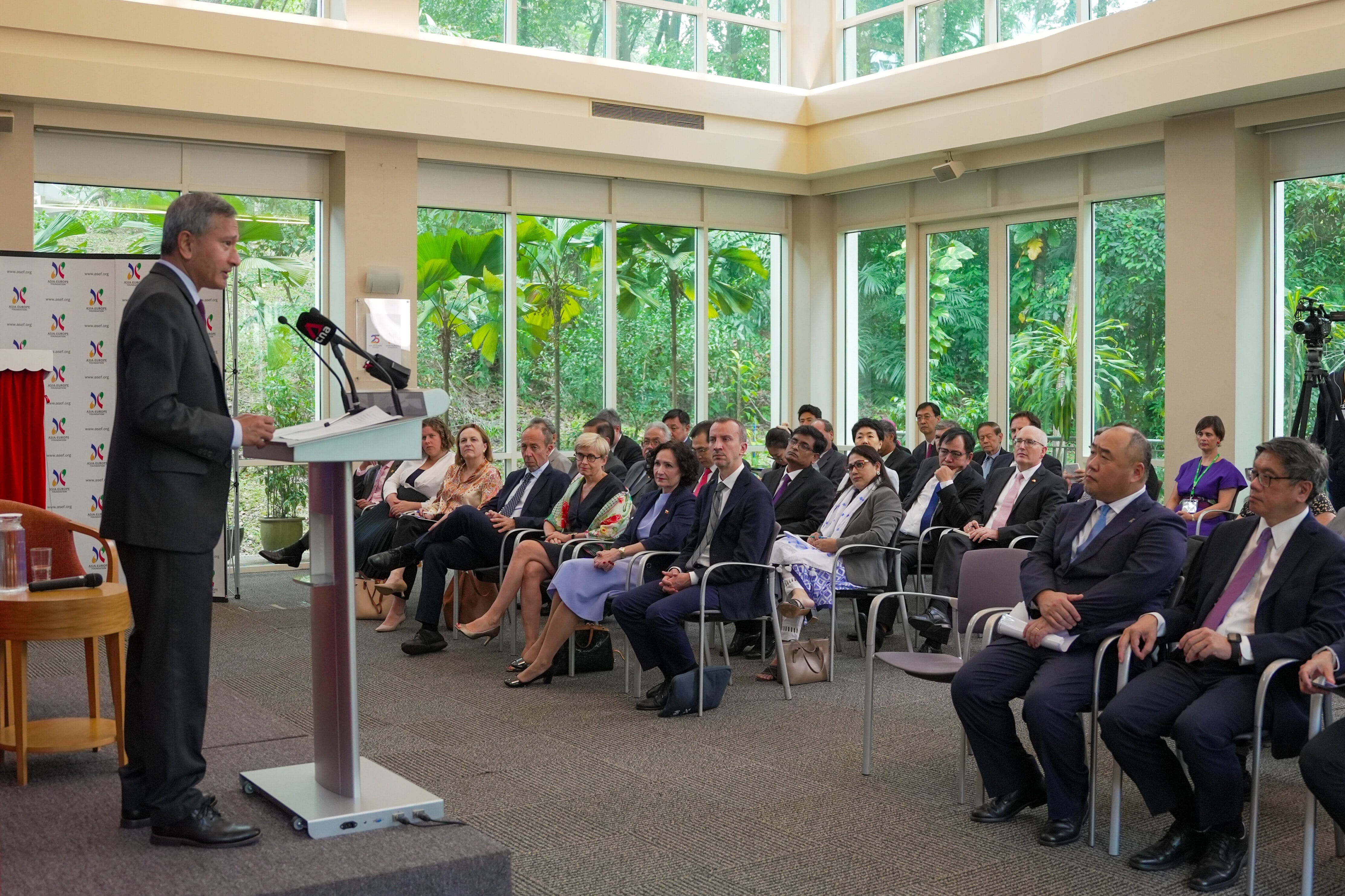 Man at podium addresses audience in suits; lush greenery visible through large windows.